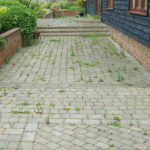 Block paved path with weeds growing through the joints beside a black clad building